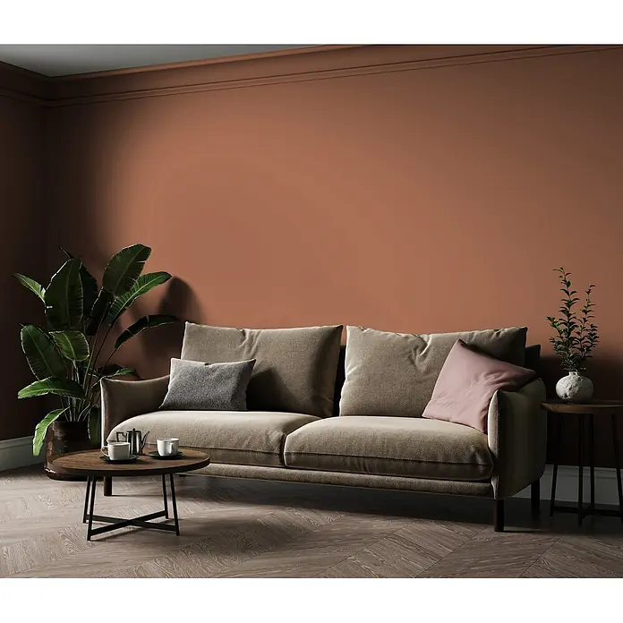 Livingwalls Wandfarbe Rot Dusty Orange DD126168 5l - Rot, Matt, 5 l - Ein beiges Stoffsofa mit zwei Zierkissen (grau und rosa) in einem Wohnzimmer mit Terrakotta-Wänden, einem runden Holztisch und Pflanzen.