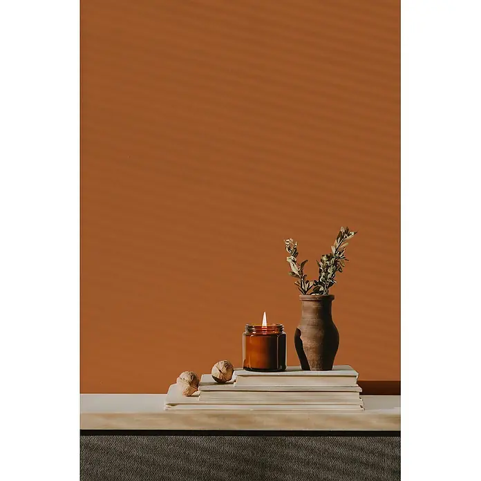 Livingwalls Wandfarbe Orange Dusty Orange DD126149 2,5l - Orange, Matt, 2,5 l - Brennende Kerze im bernsteinfarbenen Glas auf Büchern, neben einer rustikalen Vase mit Grünzeug und Walnüssen, vor orangefarbener Wand.