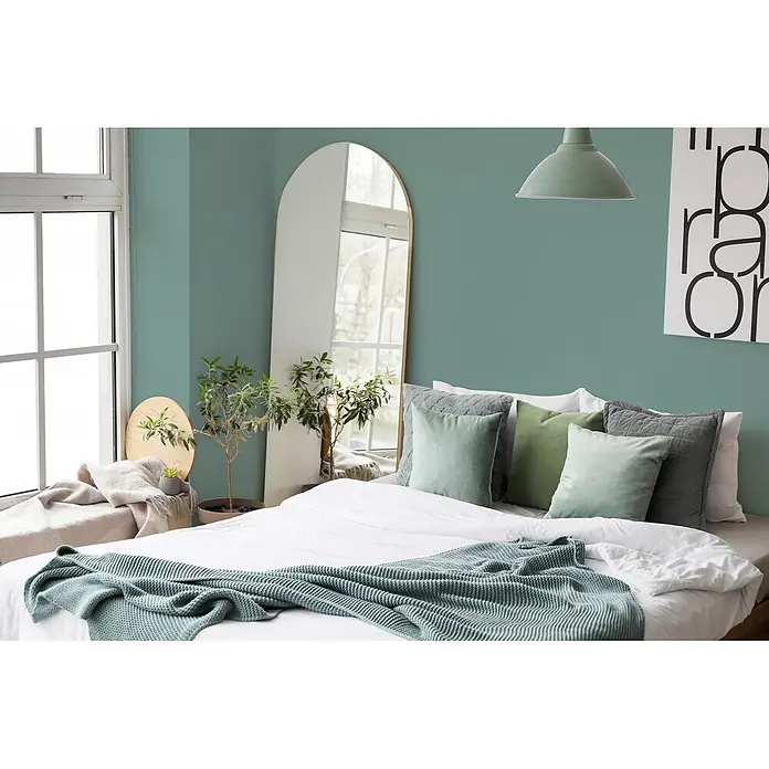 Livingwalls Wandfarbe Grün Soft Green DD125928 5l - Grün, Matt, 5 l - Ein Bett mit weißer Bettdecke, grünen und grauen Kissen sowie einer grünen Strickdecke, die ein gemütliches Schlafzimmerambiente schaffen.