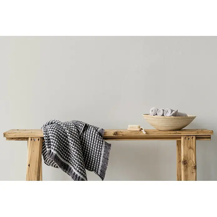 Livingwalls Wandfarbe Creme Shady Sugar 1008 2,5l - Matt, 2,5 l - Ein dunkelgrau-weißes Waffeltuch liegt über einer rustikalen Holzbank, daneben eine Holzschale mit gerollten Handtüchern und einer Bürste.