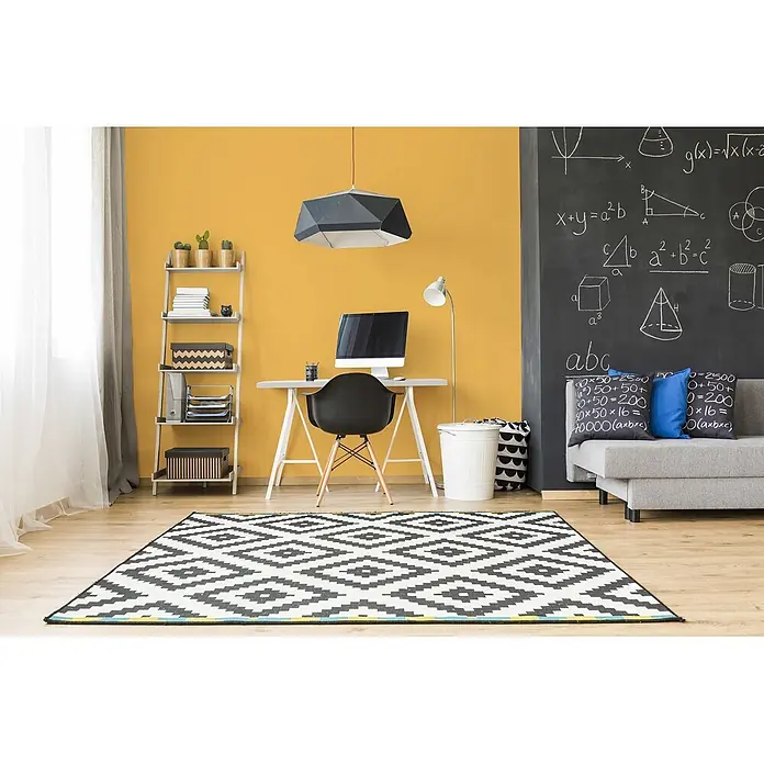 Livingwalls Wandfarbe Gelb Sunny Yellow DD126111 5l - Gelb, Matt, 5 l - Modernes Arbeitszimmer mit gelber Wand, Kreidetafel, grauem Schreibtisch, schwarzem Stuhl und einem markanten schwarz-weißen Geometrie-Teppich auf Holzfußboden.