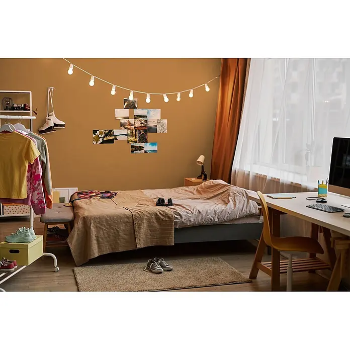 Livingwalls Wandfarbe Orange Smooth Saffron DD126136 1l - Orange, Matt, 1 l - Ein gemütlich eingerichtetes Schlafzimmer mit Bett, neutraler Bettwäsche, Wandgalerie, Lichterkette und Schreibtisch mit Computer.