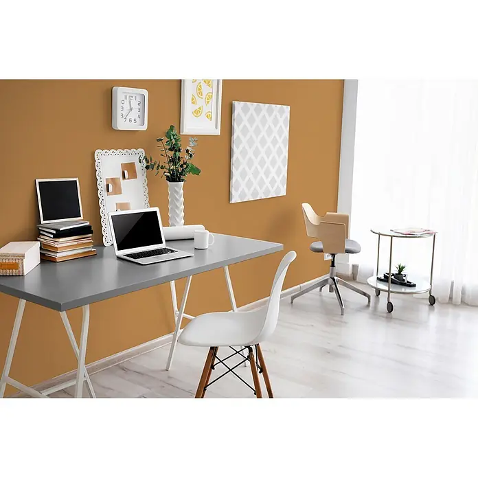 Livingwalls Wandfarbe Orange Salted Caramel 5007 2,5l - Matt, 2,5 l - Ein modernes Heimbüro mit grauem Schreibtisch, Laptop, Büchern und einem weißen Stuhl mit Holzbeinen vor einer warmen Senfwand.