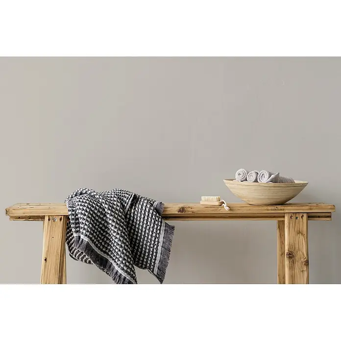Livingwalls Wandfarbe Taupe Opened Oyster 1016 2,5l - Matt, 2,5 l - Spa-Utensilien auf einer Holzbank: ein graues gemustertes Handtuch, eine Holzkörperbürste und eine Schale mit gerollten hellgrauen Handtüchern.