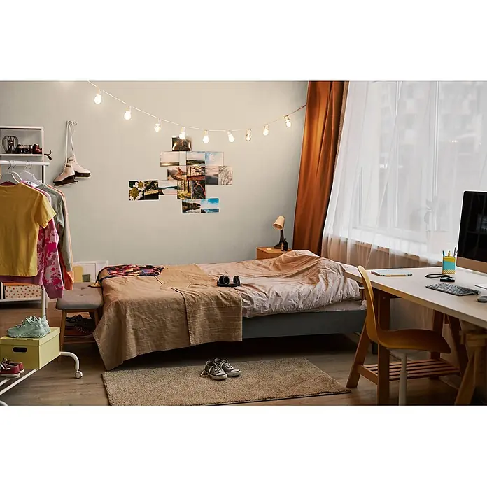 Livingwalls Wandfarbe Beige Mellow Brown DD126028 1l - Beige, Matt, 1 l - Gemütliches Schlafzimmer mit gemachtem Bett, Schreibtisch, Kleiderständer. Lichterkette und Fotos schmücken die Wand.