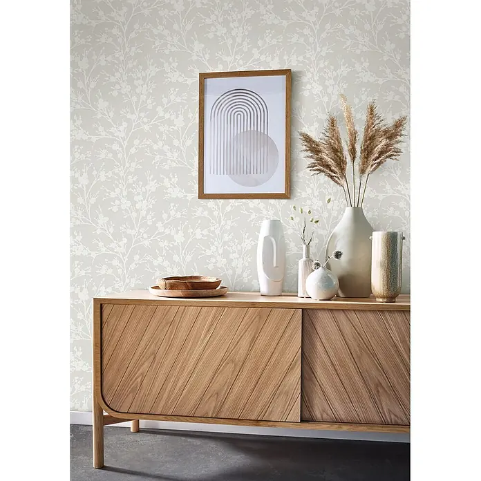 Livingwalls Vliestapete Floral, greige - 10.050 x 530 mm - Helles Holz-Sideboard mit diagonalen Lamellentüren, dekoriert mit Vasen, Kunst und Pampasgras, vor gemusterter Tapete.