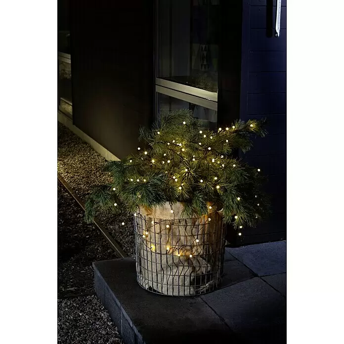 Konstsmide LED-Lichterkette Globes - Außen, Lichtfarbe: Warmweiß, Anzahl Leuchtmittel: 40 Stk., Batteriebetrieben - Eine getopfte immergrüne Pflanze mit warmweißen Lichterketten in einem Drahtgeflecht- und Sackleinenbehälter, schmückt nachts einen Außenbereich.
