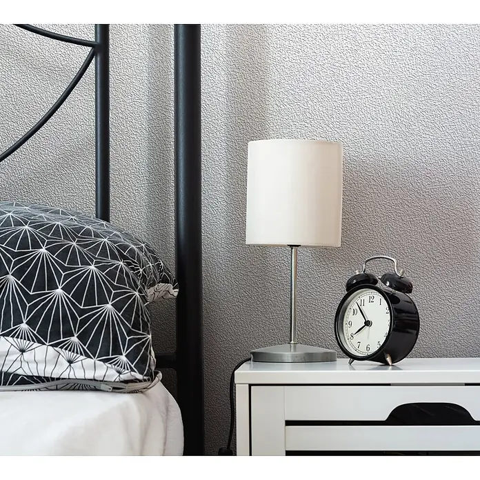 Livingwalls Vliestapete Struktur, weiß -  - Weißer Nachttisch mit Lamellen-Design und Griffaussparungen, darauf eine Lampe und Wecker, neben einem Bett mit gemustertem Kissen.