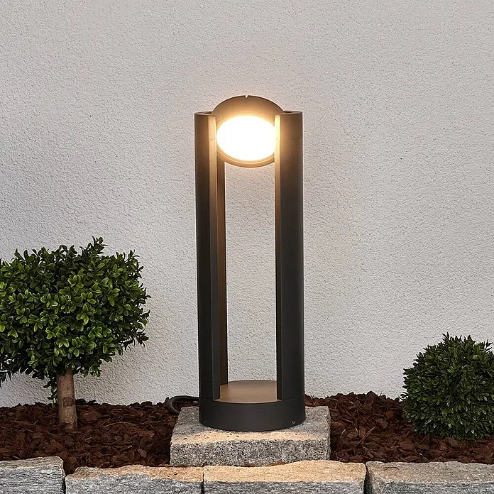 Lucande Außensockelleuchte Dylen -  - Moderne schwarze Gartenwegleuchte aus Metall mit leuchtender Milchglaskugel, beleuchtet einen Gartenbereich mit Pflanzen und Mulch.