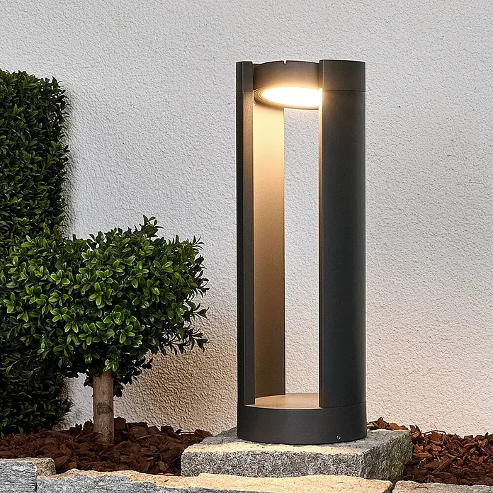 Lucande Außensockelleuchte Dylen -  - Dunkelgraue zylindrische Pollerleuchte für außen mit warmweißer LED-Beleuchtung, auf Steinplatte neben Pflanze stehend.