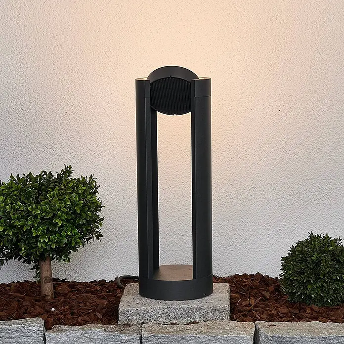 Lucande Außensockelleuchte Dylen -  - Schwarze Außenpollerleuchte auf Steinsockel, beleuchtet eine strukturierte Wand und den Gartenbereich.