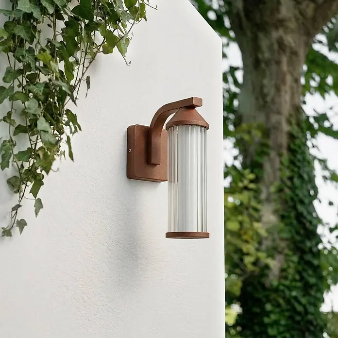 Lindby LED Wandleuchte aussen in rost, transparent, Aussenlampe Wand -  - Rostfarbene Außenwandleuchte mit geripptem Glasdiffusor neben grünem Efeu.