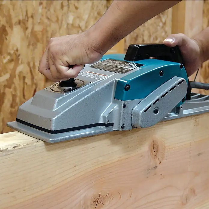 Makita Elektrohobel 1806B - 1.200 W, Hobeltiefe: 0 mm - 2 mm, Leerlaufdrehzahl: 15.000 U/min - Eine Person bedient einen blau-silbernen Makita Elektrohobel auf einem Holzbalken.