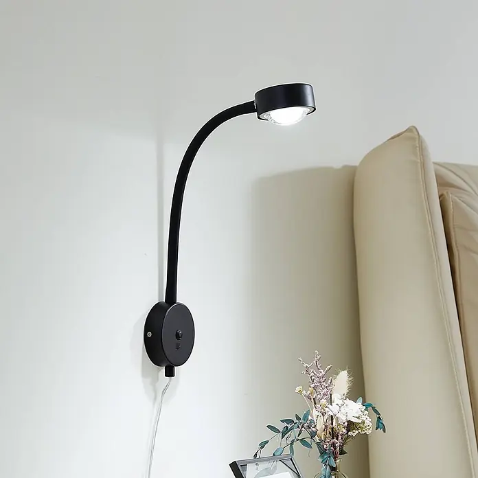Lindby Wandleuchte Jyla - L x B x H: 26,5 x 8 x 25 cm, schwarz, schwarz, 2GX53 - Schwarze, wandmontierte LED-Leselampe mit flexiblem Schwanenhals, leuchtet eine Wand neben einem cremefarbenen Kopfteil an.