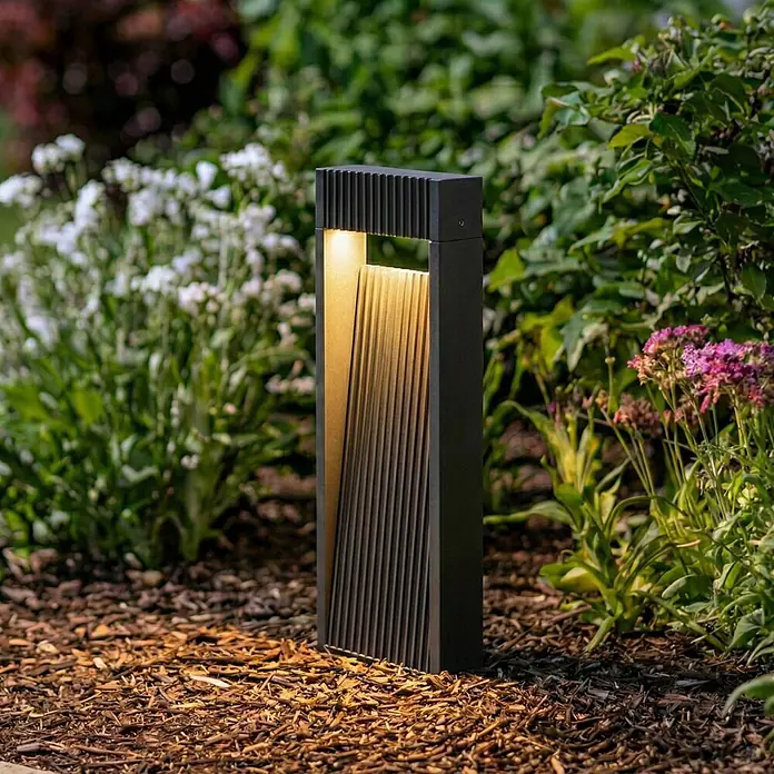 Lucande Außensockelleuchte Tavion -  - Schwarze moderne Pollerleuchte mit gerippter Innenfläche und warmer Beleuchtung, die Gartenmulch erhellt.