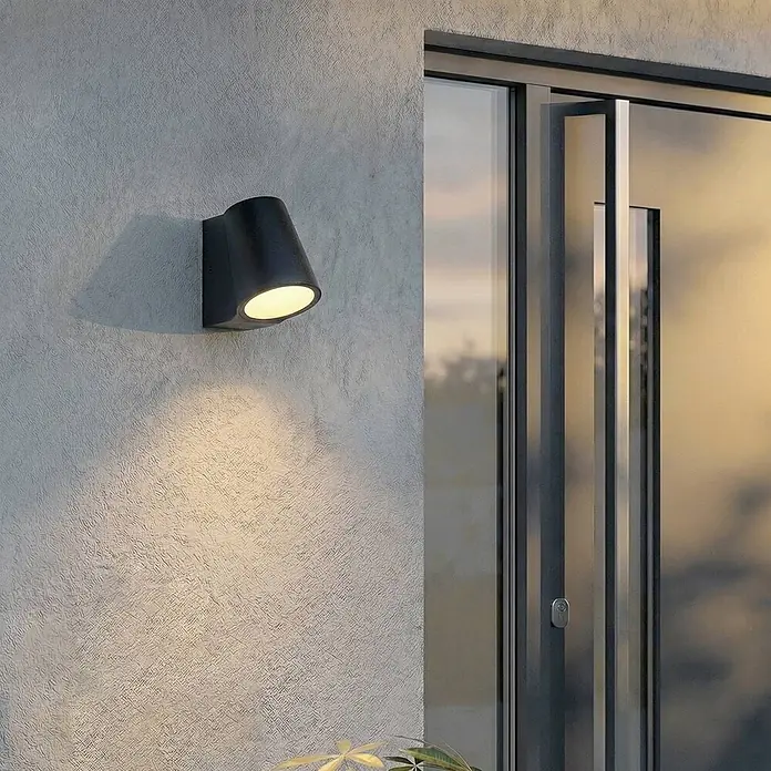 Lindby Außenwandleuchte Illuvia - schwarz, schwarz, IP44 - Schwarze Wandleuchte beleuchtet eine strukturierte Wand neben einer modernen Glastür.