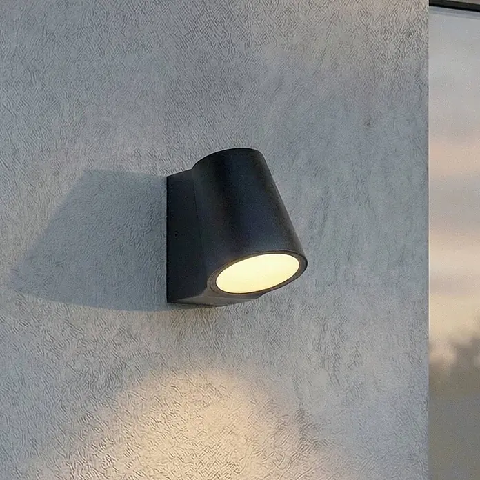 Lindby Außenwandleuchte Illuvia - schwarz, schwarz, IP44 - Schwarze Wandleuchte beleuchtet eine strukturierte Wand mit warmem Licht.