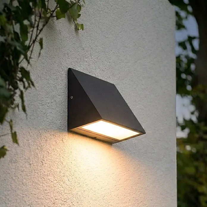 Lindby Außenwandleuchte Triava - schwarz, IP65 - Schwarze, geometrische Außenwandleuchte, die warmes Licht nach unten auf eine strukturierte Wand wirft.