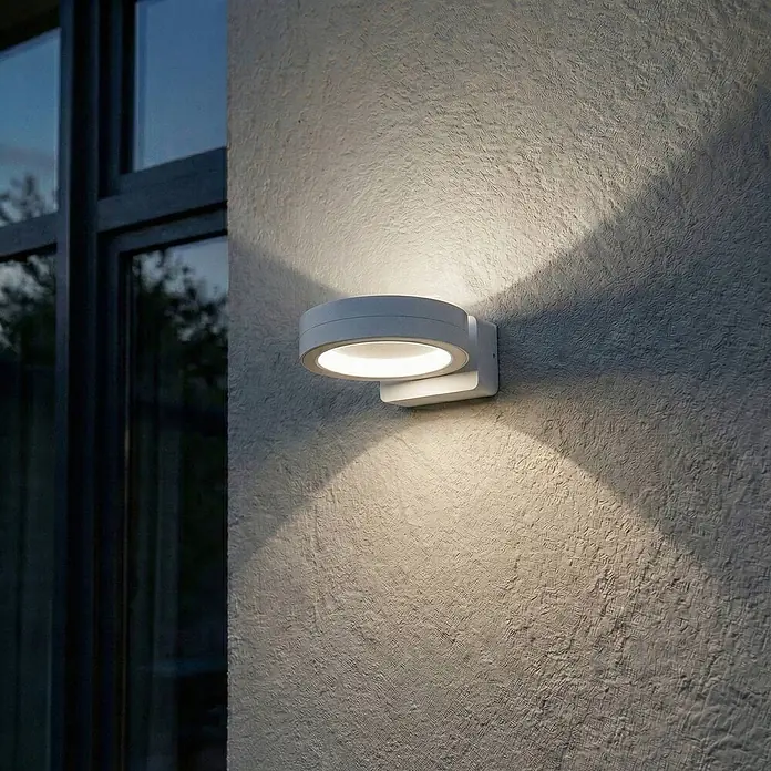 Lindby Außenwandleuchte Selviro - 24,3 x 20 x 10 cm, weiß / opal, weiß / opal, IP65 - Runde weiße Außenwandleuchte an einer strukturierten Wand, die warmes Licht nach oben und unten abgibt, neben einem Fenster.