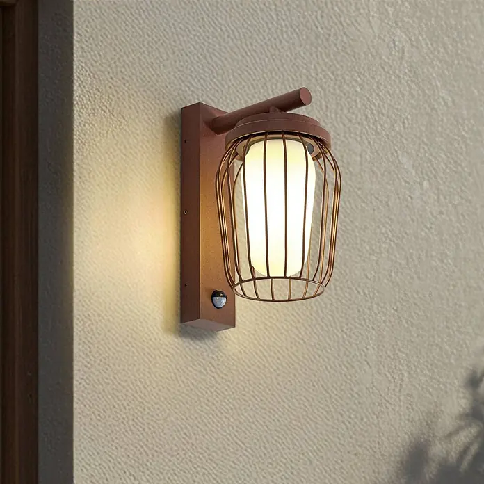 Lucande Außenwandleuchte Chandan - 23,4 x 16,5 x 30 cm, braun / rost, IP44 - Bronzefarbene Außenwandleuchte mit Käfigdesign, mattiertem Diffusor und integriertem Bewegungssensor, an einer Außenwand montiert.