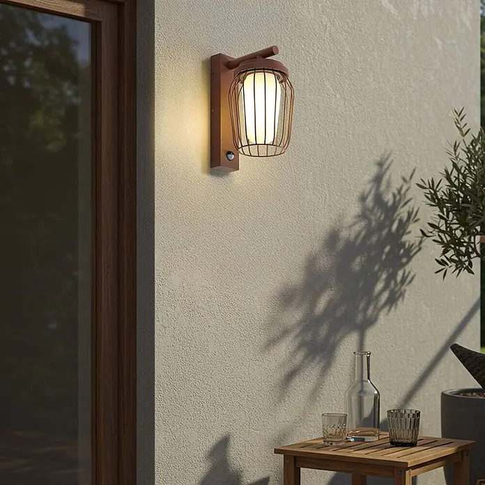 Lucande Außenwandleuchte Chandan - 23,4 x 16,5 x 30 cm, braun / rost, IP44 - Außenwandleuchte mit dunklem Holzsockel und Metallkäfigdesign, die warmes Licht auf eine strukturierte Wand strahlt.