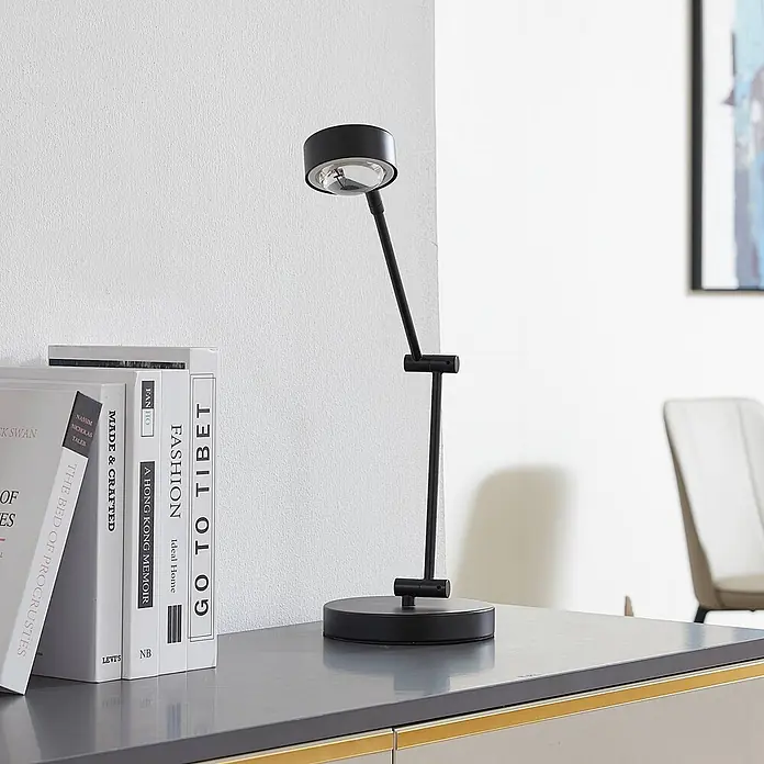 Lindby Tischleuchte Jyla - schwarz, schwarz - Schwarze, verstellbare Schreibtischlampe aus Metall mit rundem Fuß, Kopf und Gelenkarm, auf einem Schreibtisch neben Büchern.