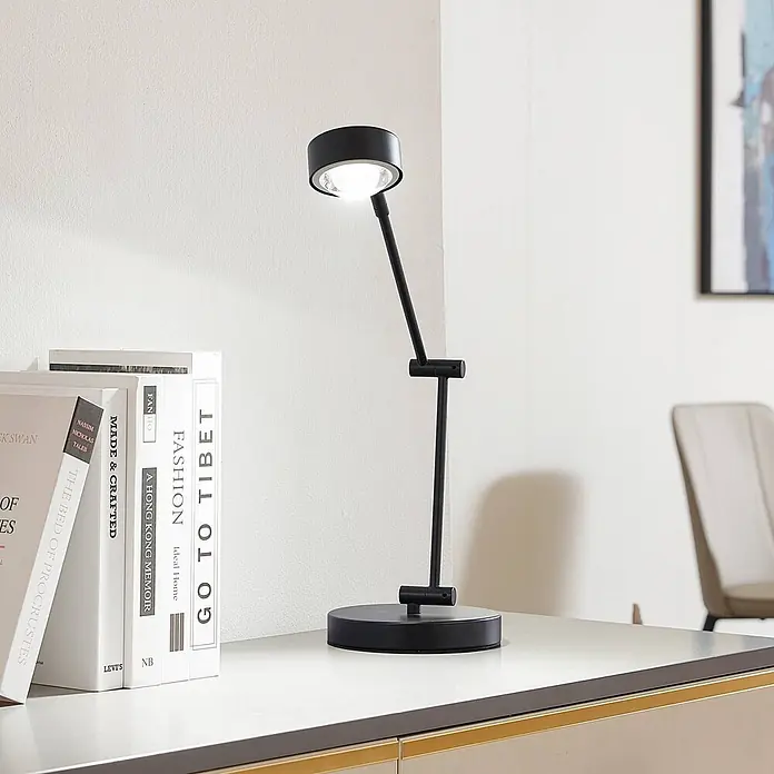 Lindby Tischleuchte Jyla - schwarz, schwarz - Verstellbare schwarze Schreibtischlampe mit rundem Fuß und Kopf, beleuchtet einen Schreibtisch neben Büchern.