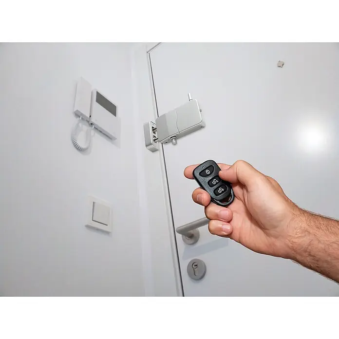 Arregui Cerradura electrónica CI10 - Plateado, Apto para: Puertas, Señal de alarma: 110 dB - Una mano usa un mando a distancia negro para operar un sistema de cerradura inteligente blanco y plateado instalado en una puerta.