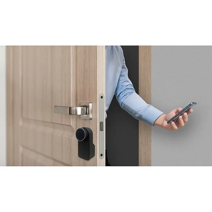 Arregui Cerradura electrónica Spin - Mediante app - Cerradura inteligente negra en una puerta de madera clara, operada por una persona con un smartphone.