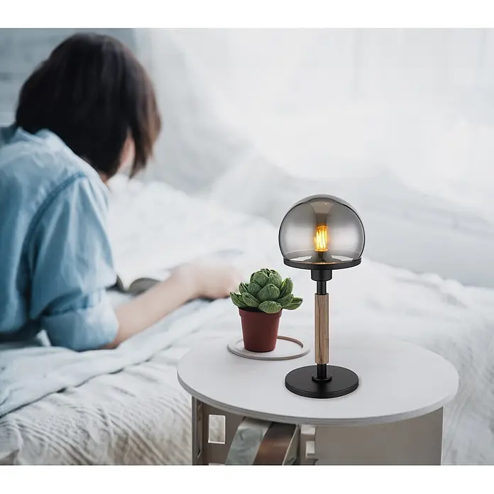Globo Tischleuchte Haku - 40 W, Höhe: 400 mm, E27 - Moderne Tischlampe mit Rauchglasschirm und Holzelement auf einem Beistelltisch, neben einer lesenden Person.