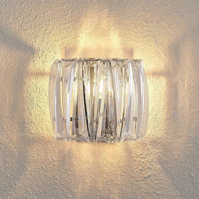 Lindby Wandleuchte Sofia - L x B x H: 14 x 23 x 20 cm, klar / transparent, E14 - Elegante Kristall-Wandleuchte, die warmes Licht auf eine strukturierte Wand wirft.