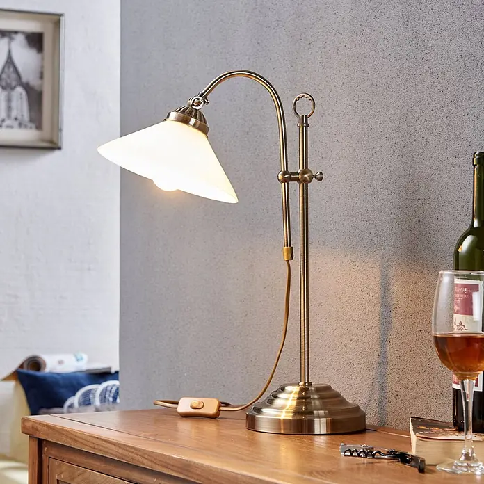 Lindby Tischleuchte Otis - bronze / altmessing, E14 - Verstellbare Tischleuchte aus antikem Messing mit weißem, konischem Glasschirm, beleuchtet auf einem Holztisch.