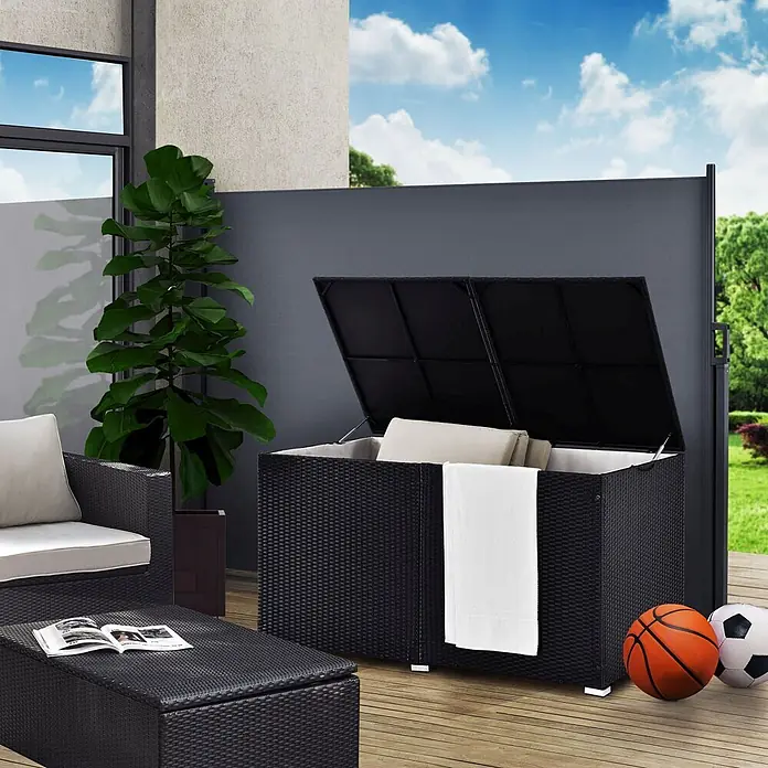 Juskys Garten-Aufbewahrungsbox Ikaria - L x B x H: 83 x 145 x 80 cm, Schwarz, Kunststoff - Schwarze Rattan-Gartenbox mit offenem Deckel, Kissen und weißem Handtuch auf einer Terrasse.