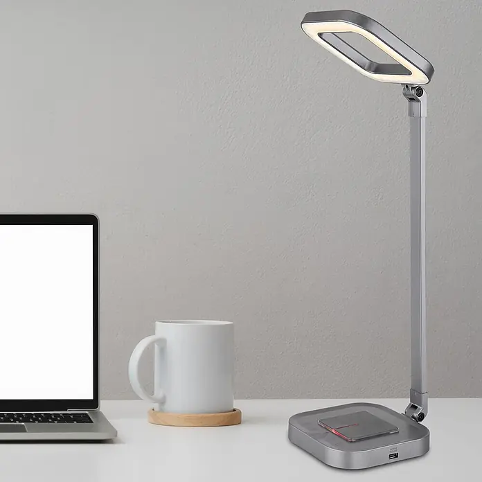 Globo LED-Tischleuchte Monaco - 8 W, L x B x H: 43 x 14,5 x 58 cm, Grau - Silberne Schreibtischlampe mit leuchtendem Rechteckkopf auf weißem Tisch. Neben Laptop und Tasse, mit kabelloser Ladefläche und USB-Anschluss.