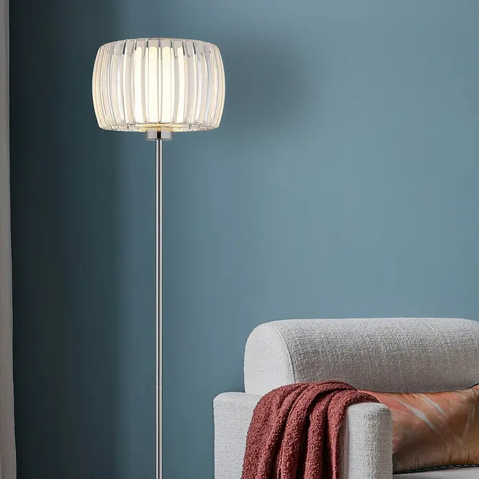 Globo Stehleuchte Maribel - 60 W, Höhe: 150 cm, E27 - Hohe Stehlampe mit klarem, geripptem Zylinderschirm und Chromfuß, beleuchtet neben einem Stoff-Sessel.