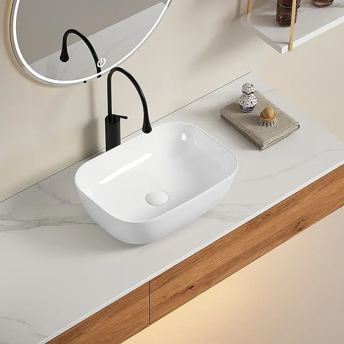 Kiamami Valentina Aufsatzwaschbecken - 330 x 460 cm, Weiß - Weißes rechteckiges Keramik-Aufsatzwaschbecken mit schwarzem Wasserhahn auf marmorierter Waschtischplatte, mit Reflexion eines beleuchteten Spiegels.