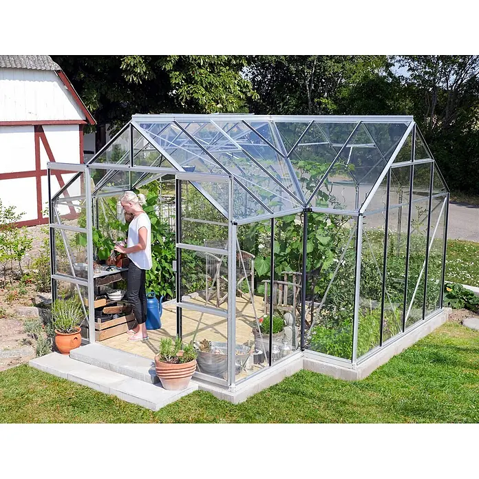 Vitavia Gewächshaus Sirona - Außenmaß inkl. Dachüberstand (B x T): 383 x 383 cm, 3 mm, Aluminium - Ein geräumiges Gewächshaus mit Glaswänden und einem silbernen Rahmen steht im Garten, eine Frau gießt Pflanzen im Inneren.