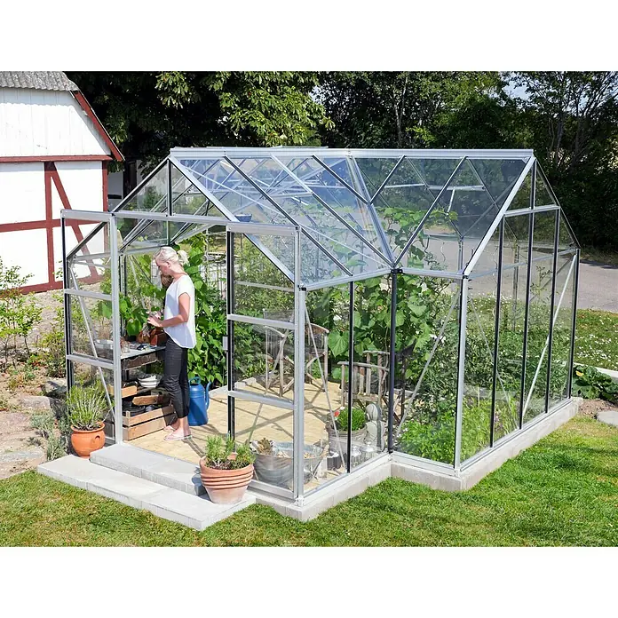Vitavia Gewächshaus Sirona - Außenmaß inkl. Dachüberstand (B x T): 383 x 383 cm, 3 mm, Aluminium - Ein geräumiges Gewächshaus mit Glaswänden und einem silbernen Rahmen steht im Garten, eine Frau gießt Pflanzen im Inneren.