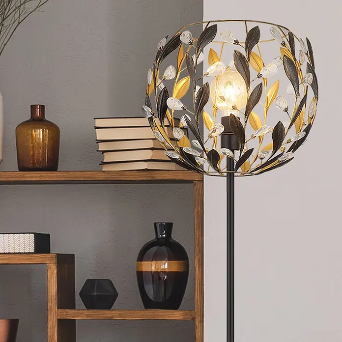 Globo Stehleuchte Mika - 40 W, Höhe: 1.570 mm, E27 - Stehleuchte mit dekorativem Schirm aus Metall- und Kristallblättern in Schwarz und Gold, beleuchtet einen Raum neben einem Holzregal.