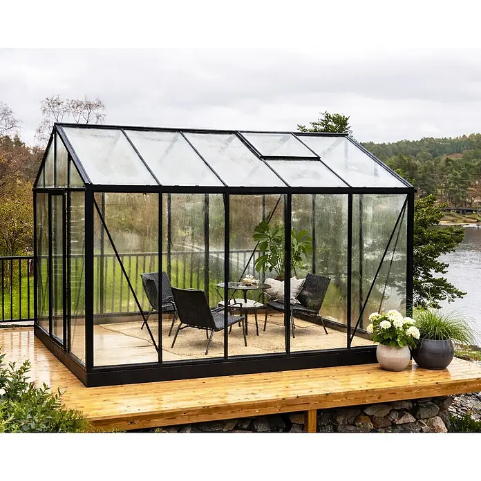 Bloomcabin Gewächshaus Aluminium Klassik - Außenmaß inkl. Dachüberstand (B x T): 235 x 384 cm, Material Seitenwand: Glas, 4 mm, Schwarz - Schwarz gerahmtes Glasgewächshaus auf Holzterrasse mit Tisch und Stühlen, Blick auf See.