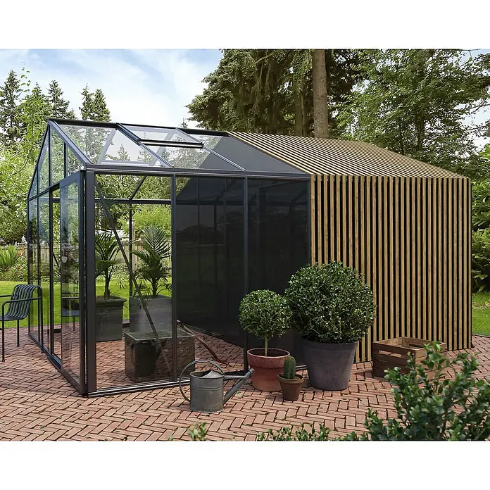 Bloomcabin Gewächshaus Aluminium Klassik mit Accoya-Fusion und großem integrierten Stauraum - Außenmaß inkl. Dachüberstand (B x T): 309 x 459 cm, Material Seitenwand: Glas, 4 mm, Schwarz - Ein modernes Gewächshaus mit schwarzem Metallrahmen und Klarglas, kombiniert mit einem Holzlamellen-Schuppen, auf einer Ziegelterrasse im Garten.