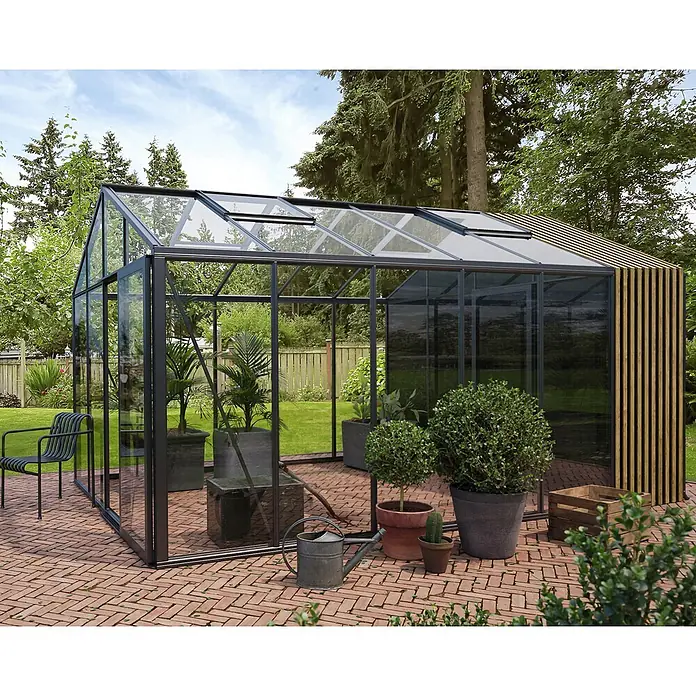 Bloomcabin Gewächshaus Aluminium Klassik mit Accoya-Fusion und kleinem integrierten Stauraum - Außenmaß inkl. Dachüberstand (B x T): 309 x 459 cm, Material Seitenwand: Glas, 4 mm, Schwarz - Großes modernes Gewächshaus mit schwarzem Rahmen, klaren Glaspaneelen und Holzlamellenwand auf Ziegelterrasse im Garten.