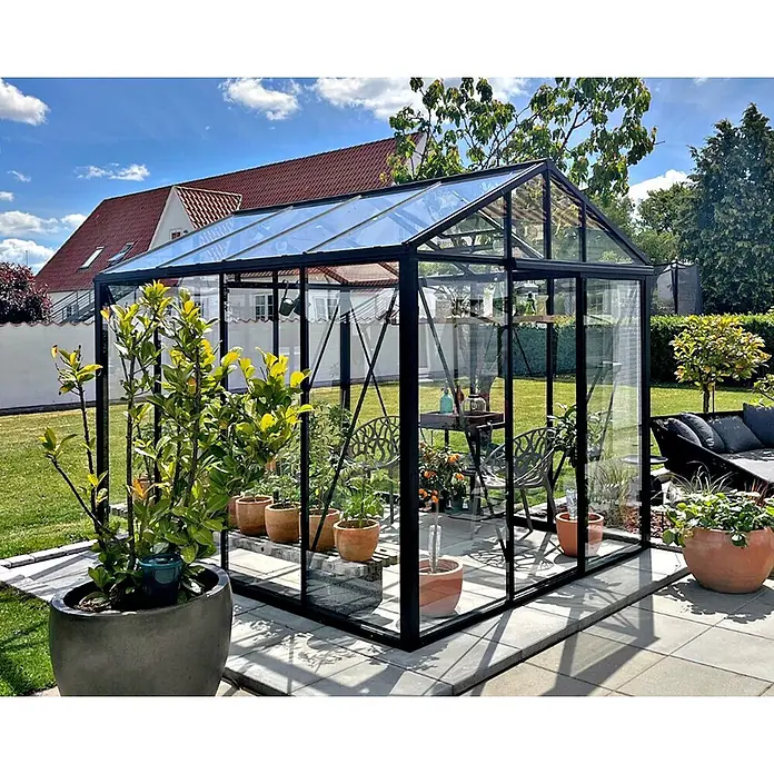 Bloomcabin Gewächshaus Aluminium Klassik - Außenmaß inkl. Dachüberstand (B x T): 235 x 309 cm, Material Seitenwand: Glas, 4 mm, Schwarz - Ein schwarz gerahmtes Gewächshaus aus Glas in einem sonnigen Garten, gefüllt mit Pflanzen und einem Sitzbereich.