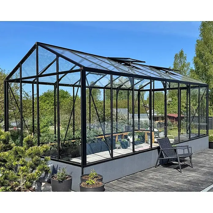 Bloomcabin Gewächshaus Aluminium für die Wandbasis - Außenmaß inkl. Dachüberstand (B x T): 309 x 608 cm, Material Seitenwand: Glas, 4 mm, Schwarz - Großes, modernes Gewächshaus mit schwarzem Rahmen, Glaspaneelen und dunkelgrauem Sockel, auf einer Holzterrasse mit Pflanzen und Stuhl.