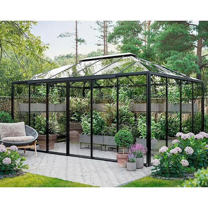 Bloomcabin Gewächshaus Aluminium Pyramide - Außenmaß inkl. Dachüberstand (B x T): 309 x 459 cm, Material Seitenwand: Glas, 4 mm, Schwarz - Großes schwarz gerahmtes Glasgewächshaus in einem Garten, gefüllt mit üppigen Pflanzen, auf einer Terrasse mit Gartenmöbeln.