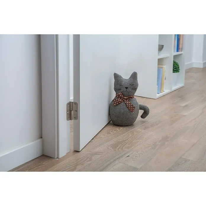 Rei Tope de puerta Gatito - Textil - Un tope de puerta de gato gris con un lazo marrón de lunares blancos sujetando una puerta abierta. Está de pie sobre un piso de madera clara.