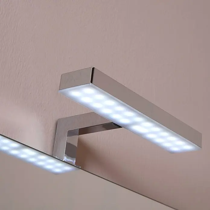 Kiamami Valentina Badmöbel-Set - Helles Holz - Verchromte, rechteckige LED-Spiegelleuchte mit vielen hellen LEDs, über einem Spiegel montiert.
