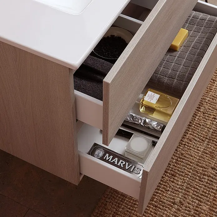 Kiamami Valentina Badmöbel-Set - Helles Holz - Badezimmermöbel aus hellem Holz mit weißer Platte und zwei offenen Schubladen mit Handtüchern und Pflegeprodukten.