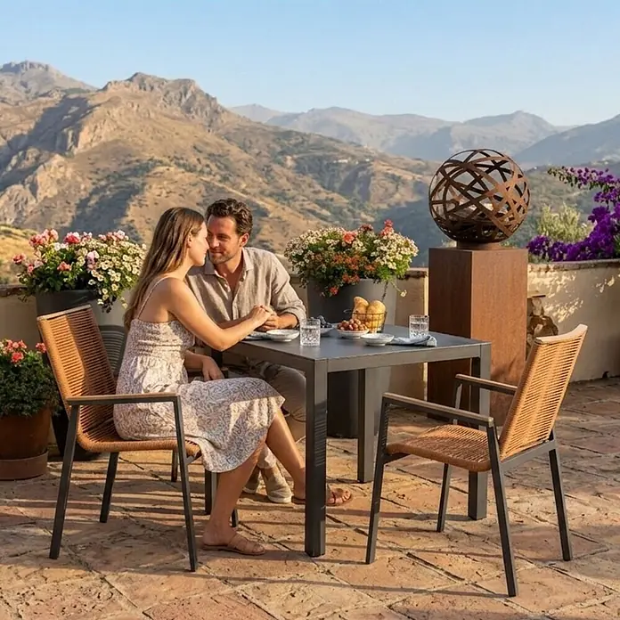 Bene Living Gartentisch-Set Merida + Hocker mit Tisch Tarifa - Gesamtumfang: 8 -tlg., Aluminium pulverbeschichtet, Textilene, Keramik, Ausziehbar - Outdoor-Essgruppe mit dunklem quadratischem Tisch und zwei Stühlen mit Naturgeflecht und dunklen Metallgestellen, von einem Paar genutzt.