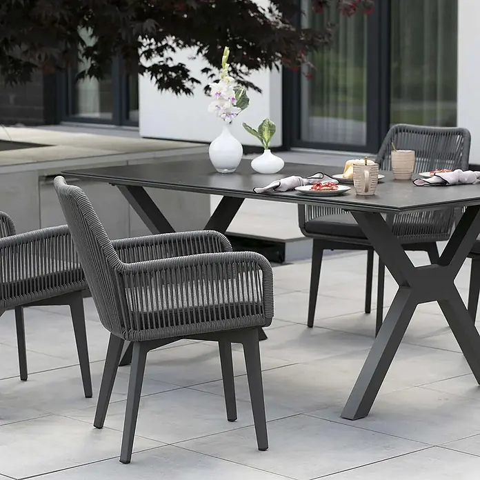 Bene Living Gartentisch-Set Marbella mit Tisch Granada - Gesamtumfang: 5 -tlg., Gestell aus pulverbeschichtetem Aluminium, Tischplatte aus gesintertem Stein (Keramik), Sitzfläche mit Rope-Bespannung (Textil/Kunststoffgemisch). - Dunkelgraue Outdoor-Essgruppe mit geflochtenen Seilstühlen und einem Tisch mit X-förmigem Fuß auf einer Terrasse.